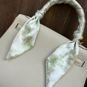 May include: Cream-colored leather handbag with a decorative silk scarf wrapped around the handle. The scarf features a white background with a green floral pattern. The bag has a textured surface and a gold-colored clasp.
