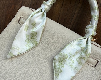 Pode incluir: Bolsa de couro creme com um lenço de seda decorativo enrolado na alça. O lenço apresenta um fundo branco com um padrão floral verde. A bolsa tem uma superfície texturizada e um fecho dourado.