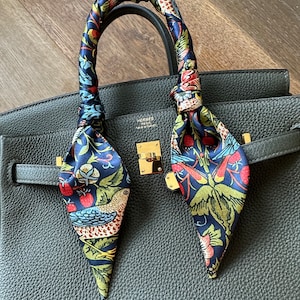 Peut inclure: Un sac à main en cuir vert foncé avec une fermeture dorée et un foulard en soie à motifs floraux noué autour des anses. Le foulard présente un fond bleu avec des fleurs et des oiseaux rouges, jaunes et verts.