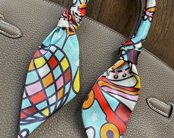 May include: A blue and white patterned silk scarf with a colorful geometric design. The scarf is tied around the handles of a brown leather handbag.