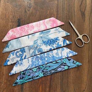 May include: Five silk scarves with various floral and botanical patterns in shades of pink, blue, and teal. A pair of small silver scissors is placed to the right of the scarves. The scarves are arranged on a dark wood surface.