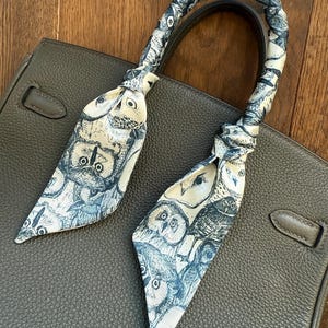 May include: A gray leather handbag with a white and blue patterned scarf tied around the handle. The scarf features a repeating pattern of owls.
