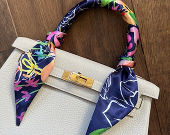 May include: A white leather handbag with a gold clasp and a colorful silk scarf tied around the handle.