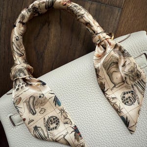 May include: A beige leather handbag with a gold clasp and a silk scarf tied around the handle. The scarf has a beige background with a black and white illustration of insects and other small creatures.