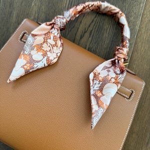 May include: A tan leather handbag with a decorative floral scarf tied around the handle. The scarf features a pattern of orange, white, and brown flowers and leaves. The bag has white stitching and a gold-colored clasp.