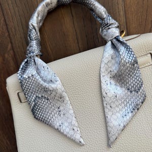 May include: A cream-coloured handbag with a textured leather surface features a handle wrapped in a silk scarf. The scarf displays a grey and white snakeskin pattern, tied securely at both ends of the handle with two long, pointed ends hanging down.