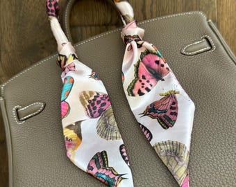 May include: A white silk scarf with a butterfly print. The scarf is tied around the handle of a brown leather handbag.
