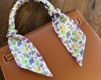 May include: A brown leather handbag with a colorful candy-printed scarf tied around the handle. The scarf features various candies in shades of yellow, orange, purple, green, and pink on a white background. The bag has a rectangular shape.