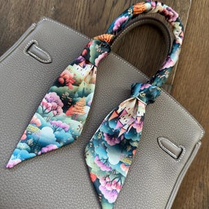 May include: A gray leather handbag with a colorful silk scarf tied around the handles. The scarf features a whimsical design of a castle, trees, and clouds.