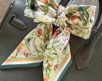 May include: A silk scarf tied in a bow around the handle of a dark grey textured leather handbag. The scarf features a botanical print with oranges, green leaves, flowers, and hummingbirds, finished with a striped border in green and orange.
