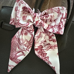May include: A white and burgundy silk scarf tied in a bow, featuring a toile de Jouy design with animal and floral motifs. The scarf is draped over a black leather handbag, adding a touch of elegance.