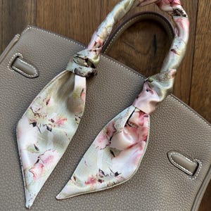 Pode incluir: Uma mala de mão de couro taupe com um lenço estampado floral enrolado na alça. O lenço apresenta flores rosa e brancas sobre um fundo verde claro. A mala tem um design estruturado com costuras brancas.