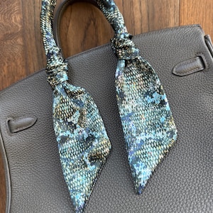 May include: A gray leather handbag with a patterned scarf tied to the handle. The scarf features a blue, green, and black design. The bag has a structured shape and a top handle.