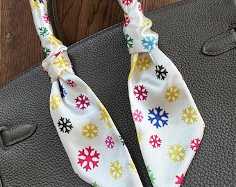 May include: A white silk scarf with a colorful snowflake pattern, tied around a black leather handbag handle. The snowflakes are in shades of red, yellow, blue, green, and pink. The handbag is a dark gray color.