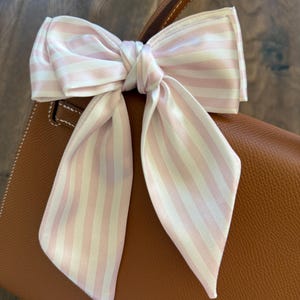 May include: A pink and white striped silk scarf tied in a bow, resting on a brown leather handbag. The scarf's stripes are evenly spaced, creating a classic and elegant look. The bow adds a decorative touch to the bag.