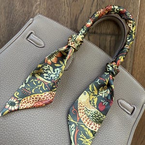 May include: A brown leather handbag with a patterned scarf tied around the handles. The scarf features a green, yellow, and red floral design with birds.