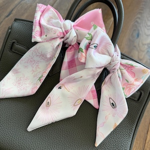 May include: Three decorative silk bow scarves in shades of pink and white are tied to the handle of a dark grey handbag. The bows feature floral, gingham, and eye-themed patterns. The handbag is made of textured leather.