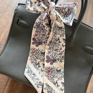 May include: A cream-colored silk scarf with a floral and butterfly pattern tied on a dark green leather handbag. The scarf features pink, purple, and green floral elements. The handbag has a structured design with a top handle.