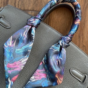 May include: A dark gray leather handbag with a colorful dragon-print scarf tied around the handle. The scarf features a repeating pattern of blue, purple, and pink dragons on a dark background. The bag has a structured design.