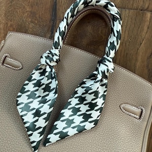 May include: A taupe leather handbag with a black and white houndstooth patterned scarf tied to the handle. The scarf adds a decorative touch to the bag. The bag has a textured surface and a top handle.