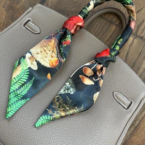 May include: A gray leather handbag with a black and green patterned scarf tied around the handle. The scarf features a design of mushrooms, ferns, and other botanical elements.