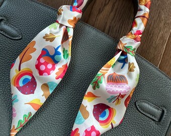 May include: A decorative silk scarf tied to a dark gray handbag handle. The scarf features a white background with colorful illustrations of acorns, leaves, and berries in shades of red, orange, blue, and green. The handbag has a textured leather appearance.
