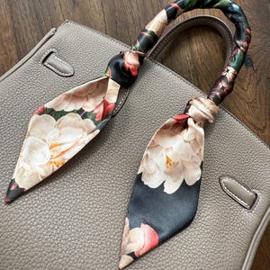 May include: A gray leather handbag with a floral patterned scarf tied around the handles. The scarf is black with pink and white flowers.