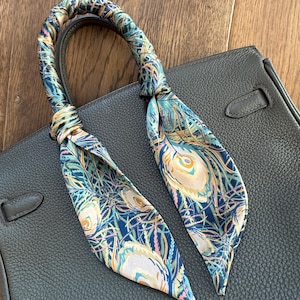 May include: A dark gray leather handbag with a decorative silk scarf tied to the handle. The scarf features a peacock feather design in shades of blue, green, and cream. The bag has a textured surface.