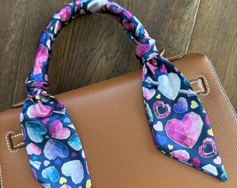 May include: A navy blue silk scarf with a heart pattern is tied to the handle of a brown leather handbag. The scarf features pink, purple, blue, and yellow heart shapes. The handbag has a gold-colored clasp.