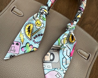 May include: A light blue silk scarf with a colorful cartoon print featuring hearts, lightning bolts, flowers, and a cassette tape. The scarf is tied around the handle of a brown leather handbag.
