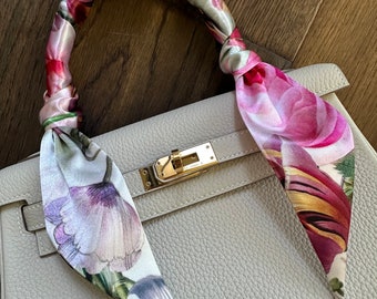 May include: A white leather handbag with a floral patterned silk scarf tied around the handle. The scarf features pink, purple, green, and yellow flowers.