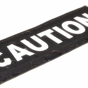 May include: Black fabric patch with the word "CAUTION" printed in white letters.