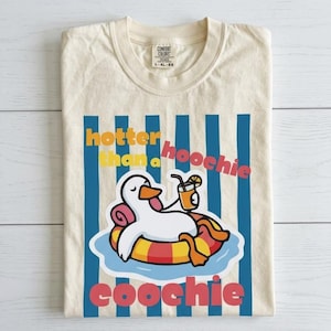 May include: Cream-colored t-shirt with a cartoon duck floating on a pool float, holding a drink. The shirt has blue vertical stripes and the text "hotter than a hoochie coochie" in orange and red.