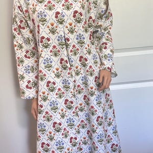 May include: A long-sleeved, floor-length dress with a floral pattern. The dress features a white background with a repeating design of red, blue, and orange flowers. The dress is displayed on a mannequin.