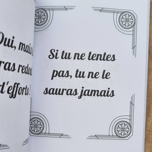 Może przedstawiać: Otwarta książka z tekstem "Si tu ne tentes pas, tu ne le sauras jamais" czarną czcionką. Białe strony mają dekoracyjne narożniki. Książka jest otwarta na drewnianej powierzchni.
