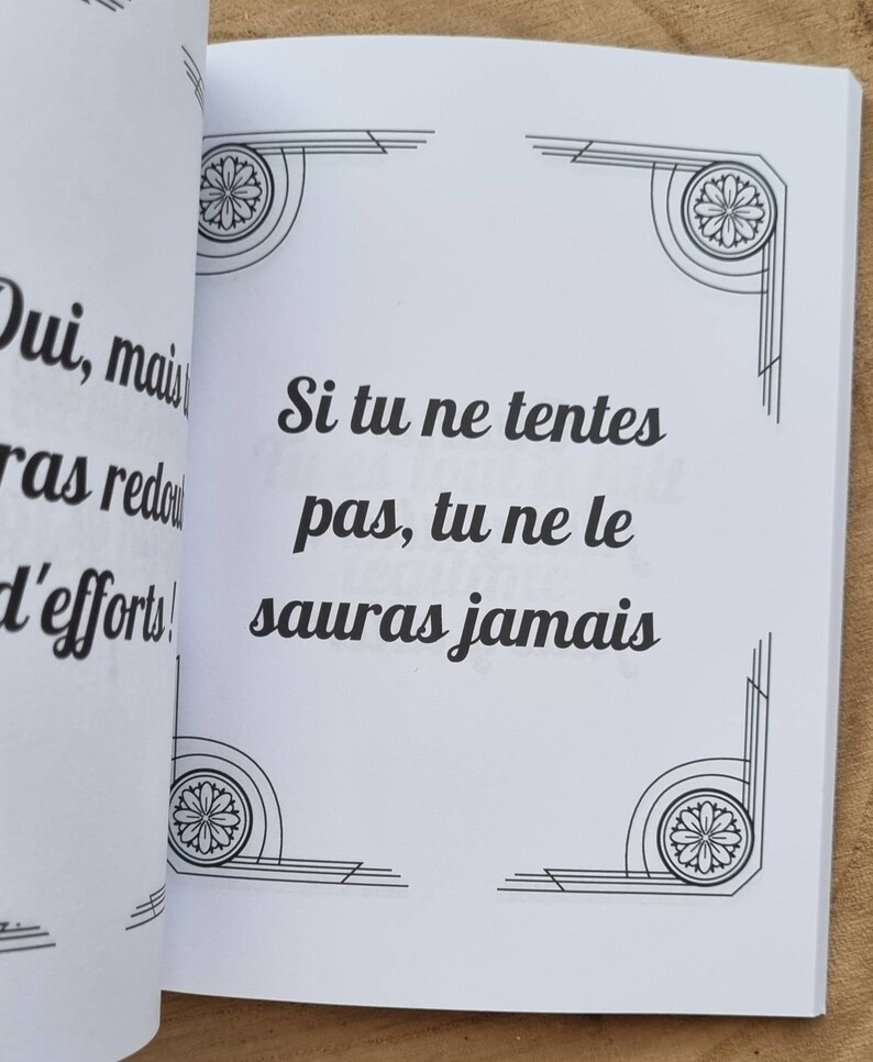 Może przedstawiać: Otwarta książka z tekstem "Si tu ne tentes pas, tu ne le sauras jamais" czarną czcionką. Białe strony mają dekoracyjne narożniki. Książka jest otwarta na drewnianej powierzchni.