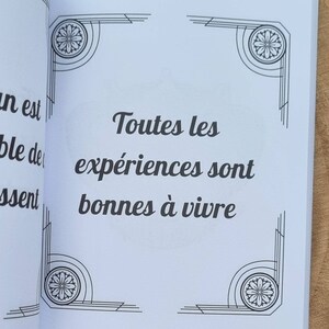 Może przedstawiać: Otwarta książka z francuskim tekstem "Toutes les exp&eacute;riences sont bonnes &agrave; vivre" w czarnym kolorze. Ozdobne narożniki z kwiatowymi i geometrycznymi wzorami obramowują tekst. Książka leży na drewnie.
