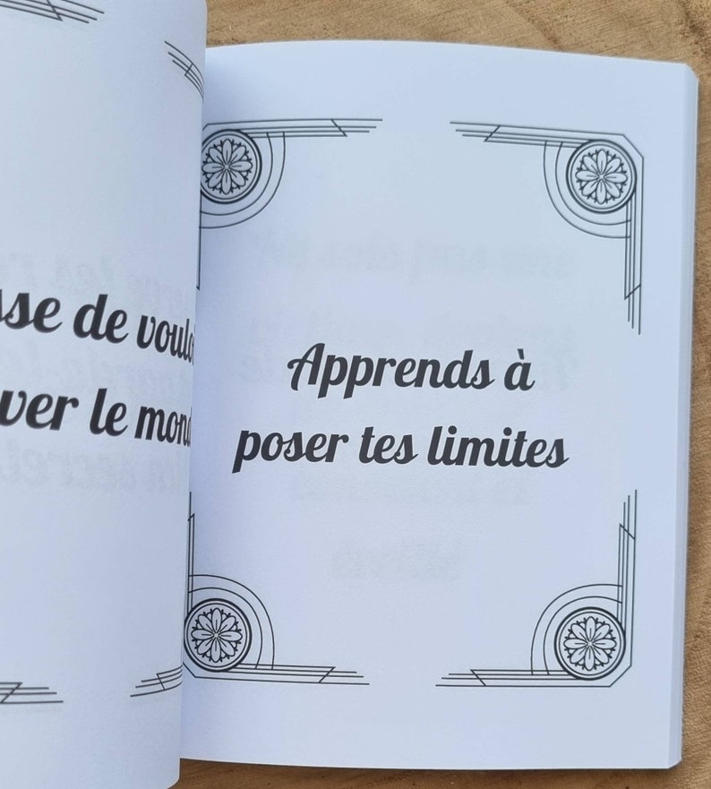 Może przedstawiać: Otwarta książka z tekstem "Apprends &agrave; poser tes limites" czcionką w kolorze czarnym. Strony są białe z dekoracyjnymi czarnymi wzorami w rogach. Książka leży na drewnianej powierzchni.