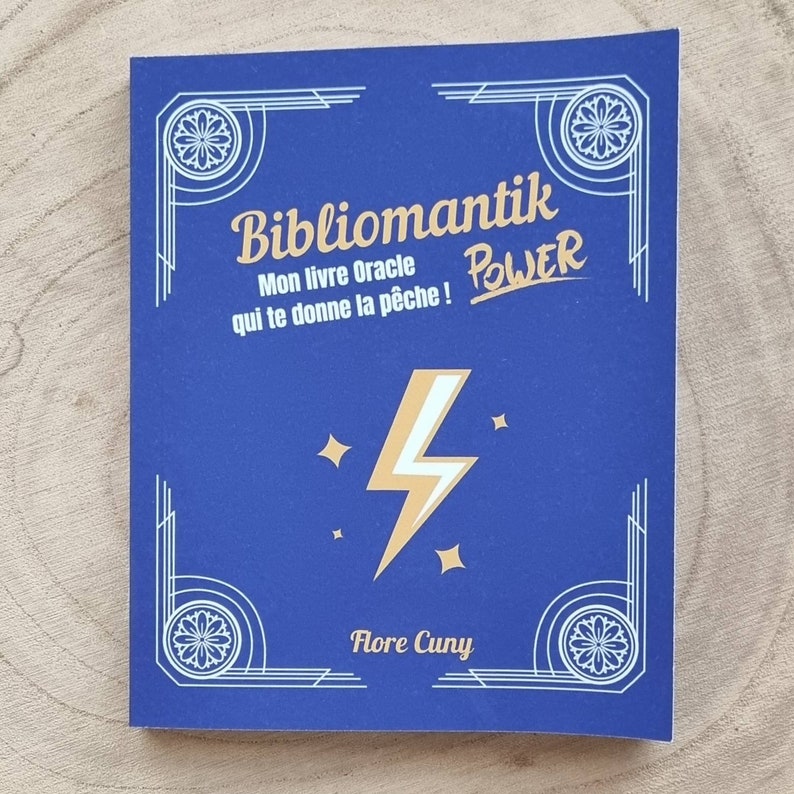 Może przedstawiać: Niebieska książka z tytułem "Bibliomantik" i słowem "POWER" w złotym kolorze. Okładka przedstawia złoty błyskawicę i gwiazdy. Nazwisko autora, Flore Cuny, znajduje się na dole. Książka ma dekoracyjne białe obramowania.