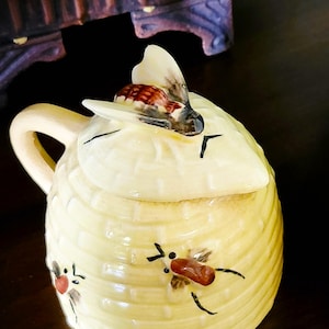 May include: A ceramic honey pot with a lid shaped like a bee. The pot is yellow with a honeycomb design and painted bees. The handle is a light cream color. The lid features a detailed bee with brown and white wings.
