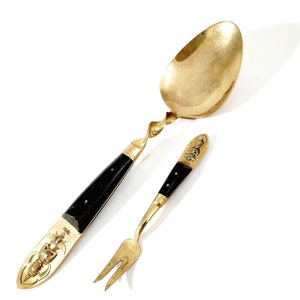 May include: A gold-toned metal spoon and a two-pronged fork featuring black handles with gold-toned accents. The spoon has a twisted neck, and both utensils display embossed figures and the word SIAM on the handle ends with black inlays.