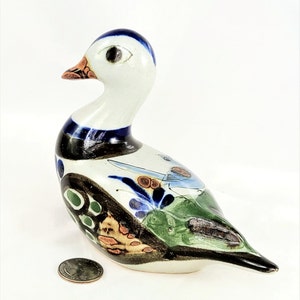 May include: A ceramic duck figurine with a white body, blue and black accents, and colorful painted details. The duck has a long neck, a small orange beak, and a detailed design on its body. A quarter is next to the duck for size comparison.
