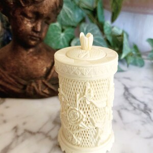May include: A cylindrical cream-colored container with a removable lid featuring a small floral finial. The exterior surface displays an intricate lattice pattern overlaid with sculpted rose blossoms and leaves, finished with a textured border.