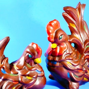 May include: A pair of ceramic rooster and hen figurines. The rooster has a red comb and tail feathers, while the hen has a smaller comb. Both are primarily brown with colorful accents of yellow, blue, and green. The background is a solid light blue.