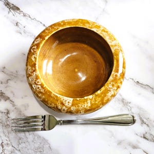 May include: A round, ceramic bowl with a light brown interior and a textured, golden-brown rim. A silver fork is positioned below the bowl on a white and gray marble surface. The bowl is empty.
