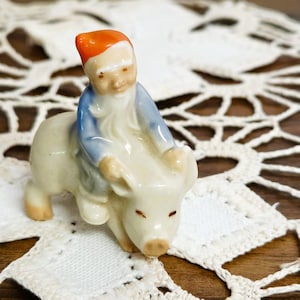 May include: A small porcelain figurine featuring a gnome with a bright orange hat and a blue jacket riding on the back of a white pig. The gnome has a long white beard and the pig has small brown painted eyes and a brown snout detail.