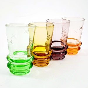 May include: Four colorful shot glasses in green, yellow, pink, and orange. Each glass has a starburst design and a unique base. The glasses are arranged in a row against a white background.