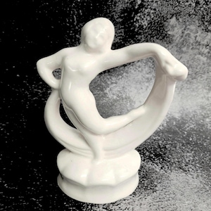 May include: A glossy white ceramic figurine depicting a stylized human figure in a dynamic pose. The figure stands on a scalloped circular base, with one arm extended and one leg bent, framed by a curved, ribbon-like element behind the body.