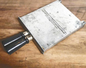 May include: Vintage silver-toned metal food slicer with two black handles. The slicer has a rectangular shape with a slightly textured surface. The text on the slicer reads: "BERNARDIN'S ADJUSTABLE-SLICER, WINCHESTER, IND. U.S.A."