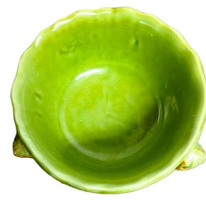May include: A bright lime green ceramic bowl with a scalloped, wavy rim and a glossy, crackled glaze finish. The bowl features three small, textured, leaf-like feet extending from the base, providing a stable foundation for the rounded vessel.